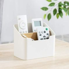 a white pen holder with remote controls in it on a wooden table next to a potted plant