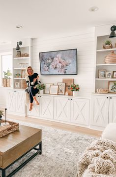 a woman sitting on the ledge of a living room with white walls and shelving