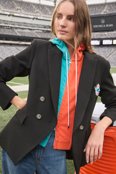 a woman in an orange shirt and black blazer stands on the field at a football stadium