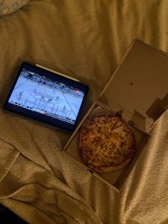 a pizza in a box next to a cell phone on a bed with a blanket