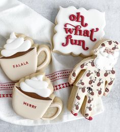 three decorated cookies and two mugs on a white cloth with red lettering that says oh what fun