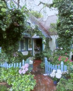 a blue house surrounded by lush green trees and flowers on a brick path leading to the front door