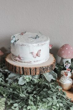 a white cake sitting on top of a wooden table next to cupcakes and plants