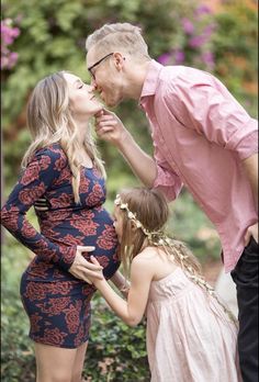 a pregnant woman kisses her husband's cheek as they stand next to their daughter