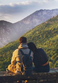 two people sitting on top of a stone wall looking at the mountains and trees in the distance