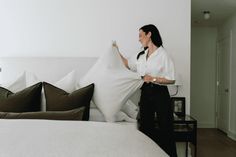 a woman standing next to a bed with pillows on it's head and holding the pillow in her hand