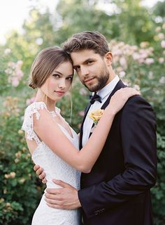 a bride and groom pose for a wedding photo in front of some bushes with pink flowers