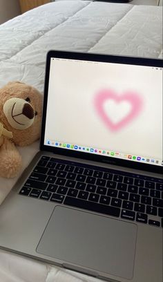 a teddy bear sitting next to a laptop on a bed with a heart drawn on the screen