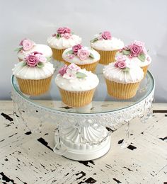 cupcakes with white frosting and pink flowers are on a glass cake plate