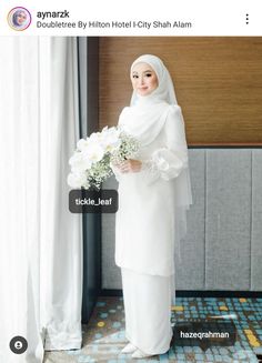 a woman standing in front of a window holding a bouquet of flowers and wearing a white hijab