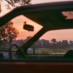 the sun is setting behind a red car with trees in the back ground and grass on the other side