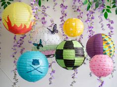 colorful paper lanterns hanging from a tree with purple and green leaves on the wall behind them