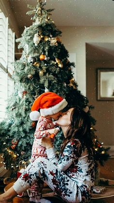 a woman sitting in front of a christmas tree kissing her baby's nose with a santa hat on