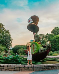 a woman wearing a hat standing in front of a fake garden with plants and flowers