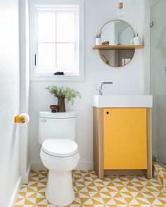 a white toilet sitting next to a yellow cabinet in a bathroom under a window with a round mirror above it