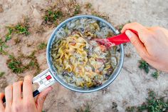 two hands holding a red spoon over a pot filled with food on the ground next to grass and dirt