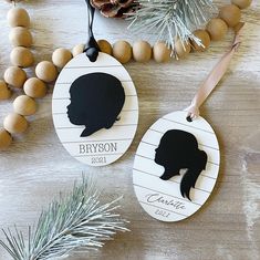 two personalized ornament hanging on a table with pine cones and evergreen branches