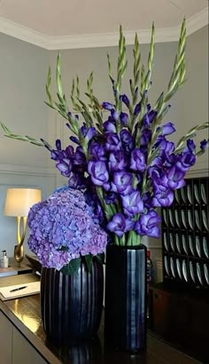 two black vases with purple flowers in them on a counter top next to a lamp