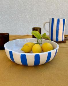 three lemons in a striped bowl on a table next to two cups and a mug