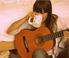 a woman sitting on a bed drinking from a cup while holding an acoustic guitar in front of her face