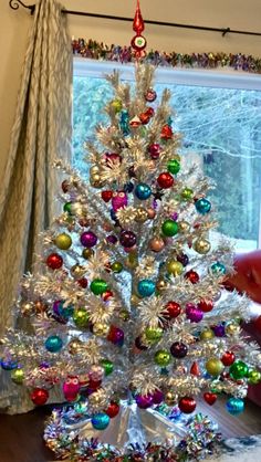 a white christmas tree in front of a window with colorful ornaments on the top and bottom