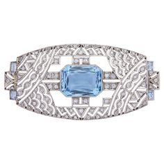 This stunning Art Deco brooch pin has been crafted from platinum and set with bright aquamarines and old cut diamonds. The central aquamarine is an emerald cut stone weighing 8.10 carats that has been rubover set. To the corners of the brooch are a further four aquamarines, these being baguette cut stones finished with a millegrain edge. The balance of the openwork brooch is set with eighty eight old cut diamonds each of H colour and SI clarity, totalling 1.50 carats. To the rear of the brooch is a yellow gold hinged pin with a platinum hook type clasp with safety catch, which are both working well and are secure. Whilst unmarked, the pin which dates to the 1920s likely originates from the United States, a perfect example of a 'Gatsby' era jewel. Whilst designed to be worn on a piece of cl Art Deco Brooch, Aqua Marine, Baguette Cut, Velvet Ribbon, Gatsby, Emerald Cut, Aquamarine, Baguette, Brooch Pin