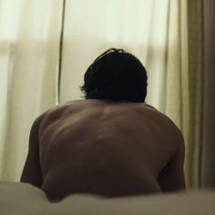 a man with no shirt sitting on a bed in front of a curtained window