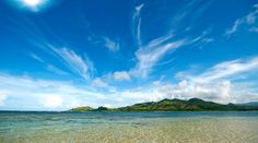 the water is crystal clear and there are clouds in the blue sky above it, with an island in the distance