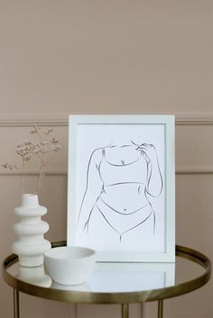 a white vase sitting on top of a table next to a framed nude woman drawing