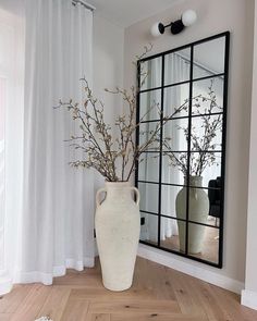 a vase with flowers in it sitting next to a large mirror on the wall near a window