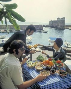 three people sitting at a table with plates of food in front of them, near the water