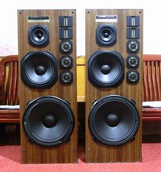 two speakers sitting side by side on top of a red carpeted floor next to a wooden bench