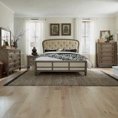 a bedroom with white walls and wooden floors, an upholstered bed, dressers, and two windows