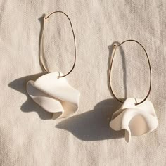 two white ceramic flowers are hanging from gold hoops on a beige fabric background, one is shaped like a flower and the other is shaped like a leaf