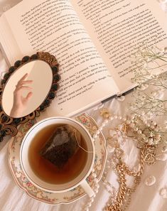 a cup of tea next to an open book on a table with jewelry and flowers