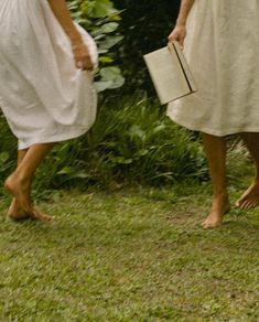 two women in white dresses are walking through the grass with an open book on their feet