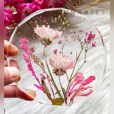 a hand holding a clear glass vase with pink flowers in it on a white furnishing