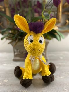 a crocheted giraffe sits in front of a vase with purple flowers