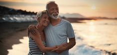 an older man and woman embracing on the beach at sunset with waves crashing in front of them