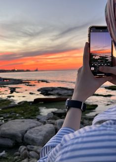 a person taking a photo with their cell phone at sunset by the water's edge