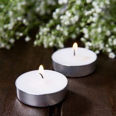 two white candles sitting next to each other on a wooden table with flowers in the background