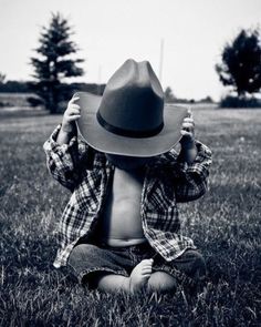 a baby boy wearing a cowboy hat sitting in the grass with his hands up to his face