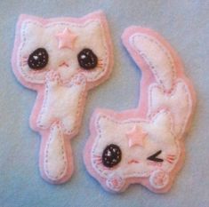 two pink and white cat brooches with black eyes on blue fabric background, one has an embellishment in the shape of a star