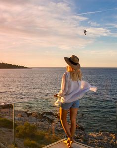 a woman standing on the edge of a cliff looking out at the ocean with a bird flying over her head