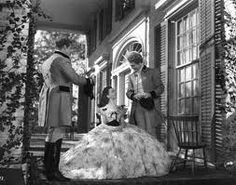 an old photo of two women in formal wear standing outside a house with flowers on the porch