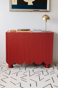 a red sideboard with books on it in front of a painting and floor rug