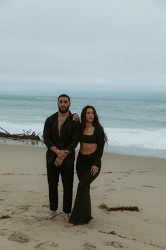 a man and woman standing on top of a sandy beach next to the ocean in black outfits