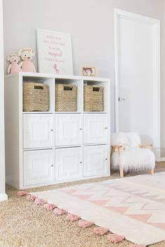 a white bookcase with baskets on top of it next to a pink rug and teddy bear