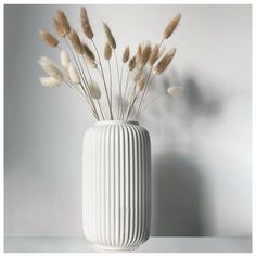 a white vase filled with dry grass on top of a table next to a wall