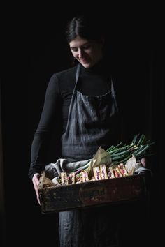 a woman in an apron holding a box full of food and vegetables on a dark background